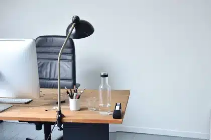 Escritorio vacío en una oficina con silla sin ocupar, reloj de pared y compañero de trabajo buscando documentos de fondo