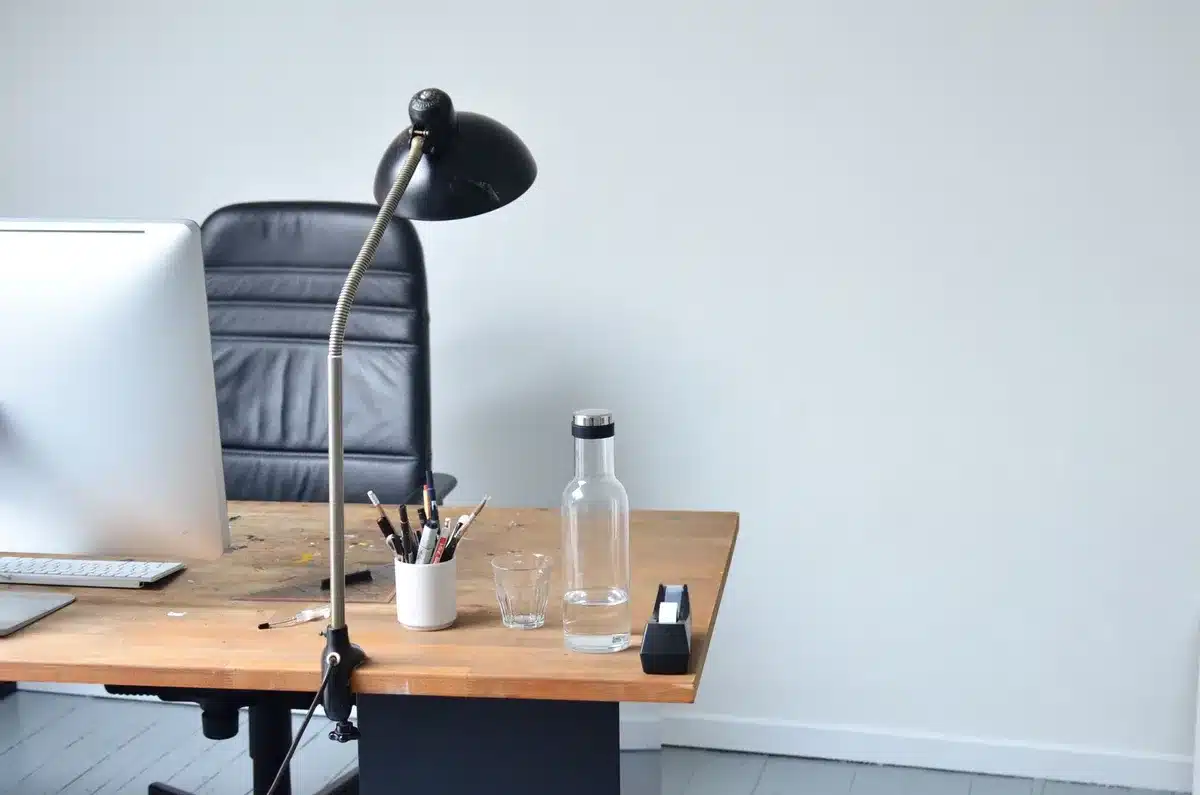 Escritorio vacío en una oficina con silla sin ocupar, reloj de pared y compañero de trabajo buscando documentos de fondo